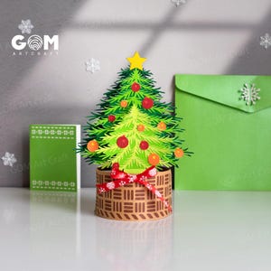 May include: A paper Christmas tree with a yellow star, red and orange baubles, and a red bow. The tree is in a brown basket with a woven pattern. A green card and envelope are in the background.