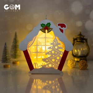 May include: A white papercraft Christmas house with a red roof and a lit window. The house has a snowy roof and a small Christmas tree inside. The house is illuminated from within, creating a warm glow.