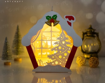 May include: A white papercraft Christmas house with a red roof and a lit window. The house has a snowy roof and a small Christmas tree inside. The house is illuminated from within, creating a warm glow.