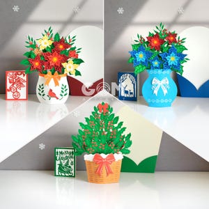 Może przedstawiać: Kolekcja trójwymiarowej sztuki papierowej o tematyce bożonarodzeniowej. Jeden przedstawia czerwono-żółtą kompozycję kwiatową w białym wazonie z motywem ptaka. Inny pokazuje niebieski wazon z niebieskimi i czerwonymi kwiatami. Trzeci to choinka w koszu.