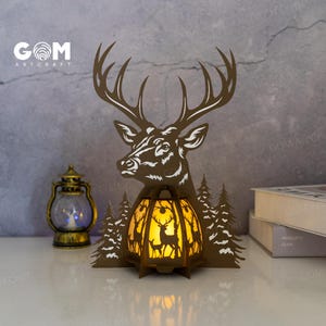 May include: A brown laser-cut deer head and lantern with the text "GOM ARTCRAFT". The lantern has a deer silhouette design and is illuminated with a warm glow. The deer head has large antlers and is surrounded by cut-out trees.