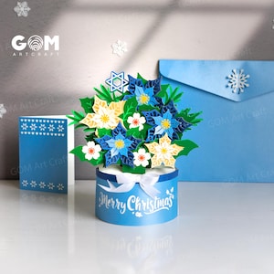 May include: A blue papercraft Christmas floral arrangement with white and yellow flowers, a Star of David, and the words "Merry Christmas." A blue card and envelope are in the background. The logo "GOM Art Craft" is visible.