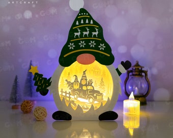 Pode incluir: Uma luz em forma de gnomo de Natal. O gnomo usa um chapéu verde com desenhos de veados e flocos de neve brancos. Ele segura uma árvore de Natal verde. A luz ilumina uma cena de gnomos em um caminhão. A marca "GOM ARTCRAFT" é visível.