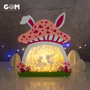 May include: A papercraft mushroom house with a fairy and a bunny inside. The mushroom is pink with white polka dots and a white and pink striped stem. The house is illuminated with a warm light.