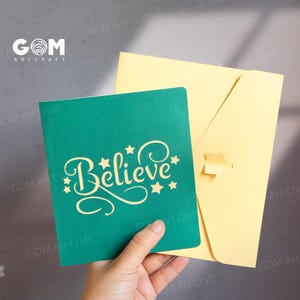 Pu&ograve; includere: Un biglietto pop-up turchese con la parola "Believe" in un carattere decorativo, impreziosito da stelle. &Egrave; accompagnato da una busta giallo chiaro. Il biglietto &egrave; tenuto in mano.