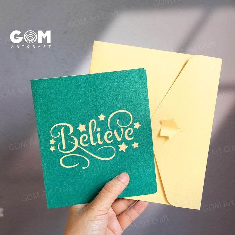 Pu&ograve; includere: Un biglietto pop-up turchese con la parola "Believe" in un carattere decorativo, impreziosito da stelle. &Egrave; accompagnato da una busta giallo chiaro. Il biglietto &egrave; tenuto in mano.