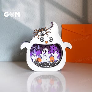 Puede incluir: Decoración de Halloween con forma de fantasma blanco con una araña en la parte superior. El interior presenta una escena espeluznante con fantasmas, calabazas y una casa embrujada. Contiene el texto "GOM Art Craft".