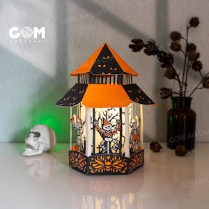 May include: A decorative Halloween lantern in orange and black. The lantern has cut-out designs of skeletons and pumpkins. The top of the lantern is orange with black detailing. The text "GOM Art Craft" is visible on the side of the lantern.