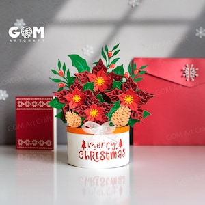 May include: A papercraft Christmas centerpiece with red poinsettias, green leaves, and pinecones in a white pot with an orange rim. The pot has "merry CHRISTMAS" text. The GOM Art Craft logo is visible.