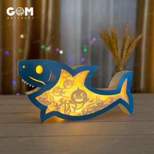 Könnte beinhalten: Eine blaue und weiße Papier-Hai-Nachtlampe mit gelbem Leuchten. Der Hai hat einen großen Mund mit scharfen Zähnen und ist von kleineren Haien und Algen umgeben. Die Lampe ist eingeschaltet und strahlt ein warmes Licht aus.
