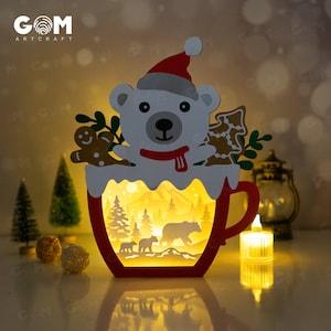 Könnte beinhalten: Ein weihnachtliches Papierkunstwerk mit einem Eisbären mit Weihnachtsmütze, einer roten Tasse und einer Winterwaldszene, die von innen beleuchtet wird. Enthält Lebkuchen- und Weihnachtsbaum-Ausschnitte.