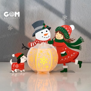 May include: A festive papercraft scene featuring a snowman, a girl, and a dog, all in winter attire. The snowman wears a hat and scarf, the girl a red coat and hat, and the dog a sweater. A glowing lantern is in the center. The text "GOM Art Craft" is visible.