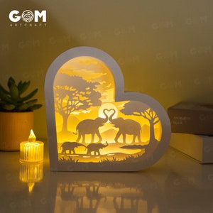 May include: A white heart-shaped lamp with a cut-out design of two elephants kissing in a savanna scene. The lamp is illuminated from within, casting a warm glow.