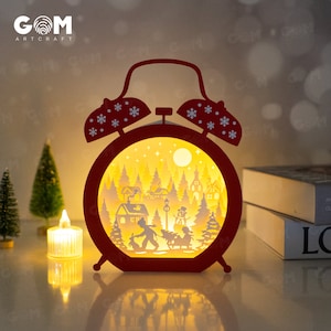 Peut inclure: Une scène de Noël illuminée en forme de réveil rouge avec un village d'hiver, un bonhomme de neige et deux enfants. La scène est éclairée par derrière, créant une lueur chaleureuse.