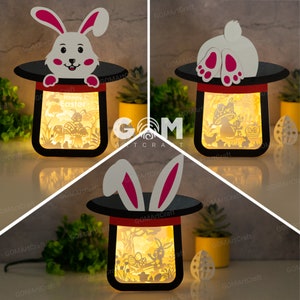 May include: Three illuminated Easter-themed papercraft night lights. Each night light features a white bunny rabbit in a black top hat with a red band. The first night light has a bunny rabbit peeking out of the top hat. The second night light has a bunny rabbit's feet sticking out of the top hat. The third night light has a bunny rabbit's ears sticking out of the top hat. Each night light has a different scene inside the top hat. The first night light has a bunny rabbit sitting on a mushroom with an egg in its paw. The second night light has a bunny rabbit holding an egg. The third night light has a bunny rabbit sitting on a mushroom with an egg in its paw.