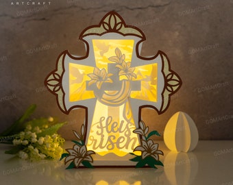 May include: A white wooden cross with a light shining through it. The cross is decorated with flowers and the words "He is risen" are written on the front. The cross is sitting on a table with a white paper egg next to it.