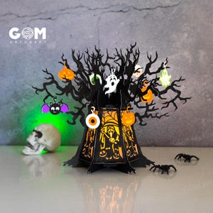 Op de afbeelding: Een zwarte lasergesneden Halloween-boomdecoratie met oranje pompoenen, geesten en een vleermuis. De boom heeft een licht aan de binnenkant en de basis heeft een spookachtig ontwerp. De schedel en spinnen dragen bij aan het Halloween-thema. De tekst "GOM ARTCRAFT" is zichtbaar.