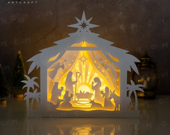 May include: A white papercraft nativity scene with a light inside, depicting the birth of Jesus. The scene includes Mary, Joseph, the baby Jesus, a donkey, a sheep, and a star. The scene is illuminated from within, creating a warm glow.