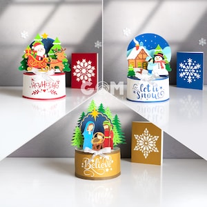 以下が含まれることがあります： クリスマスをテーマにした3つの切り絵。1つはそりに乗ったサンタクロース、もう1つは雪だるまと家、3つ目は降誕シーンです。それぞれに装飾的なカードが付いています。最初のカードには「Ho Ho Ho」、2番目のカードには「Let it Snow」、3番目のカードには「Believe」と書かれています。