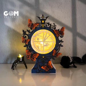 May include: A decorative, layered paper lantern with a circular design featuring a yellow moon, spiders, and bats. The lantern is adorned with orange butterflies and a black frame. A small candle holder in the shape of a hand is to the left.