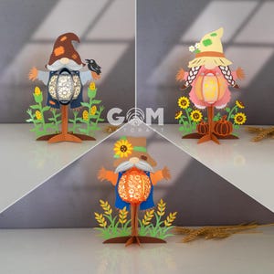 Könnte beinhalten: Drei Papierschnitt-Wichtel-Laternen mit Herbstmotiven. Einer trägt einen braunen Hut und hält einen schwarzen Vogel. Ein anderer hat einen Strohhut und Zöpfe. Der dritte hat einen Sonnenblumenhut. Jeder hält eine Laterne.