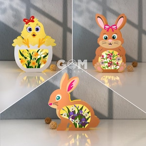 Può includere: Tre figure decorative luminose a tema pasquale: un pulcino giallo in un uovo, un coniglio marrone e un secondo coniglio marrone. Ogni figura ha un disegno floreale all'interno ed è illuminata. Il pulcino ha un fiocco rosso.