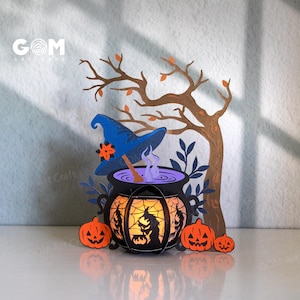 Może przedstawiać: Scena z papieru o tematyce Halloween z kotłem, dyniami i drzewem. Kocioł świeci pomarańczowym światłem, a nad nim znajduje się kapelusz czarownicy. Widoczny jest tekst "GOM Art Craft".