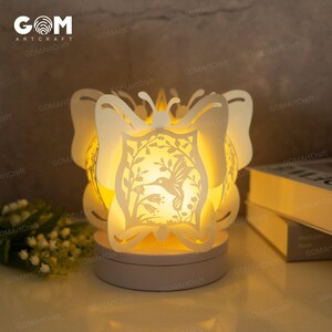 May include: A white, laser-cut butterfly lamp with a hummingbird design. The lamp is illuminated from within, casting a warm glow. The lamp is on a white base and is sitting on a table.