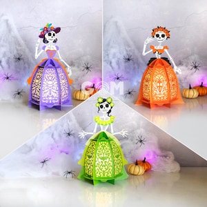 May include: Three Día de Muertos paper lanterns in purple, orange, and green, each shaped like a skeleton in a dress. The lanterns have intricate cut-out designs and are decorated with flowers. Small pumpkins and spider decorations are also present.