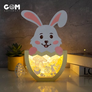 Pode incluir: Decoração de Páscoa em papel em camadas com um coelho branco saindo de um ovo rachado. A lâmpada em forma de ovo tem um brilho amarelo, iluminando uma cena com uma casa, flores e um coelho. O logotipo "GOM Artcraft" é visível.