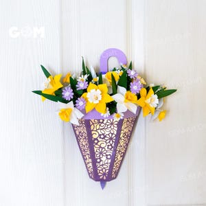May include: A purple paper lantern-shaped wall decor filled with paper flowers. The lantern has a decorative cut-out design and a handle. The flowers include yellow daffodils, white and purple flowers, and green leaves. The text "GOM Art Craft" is visible.