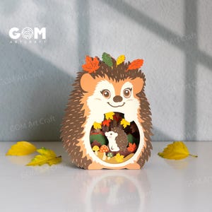 May include: A layered paper craft hedgehog decoration featuring a brown and cream colour palette with orange, yellow, and green leaf accents. The centre cutout reveals a smaller hedgehog figure surrounded by miniature mushrooms and autumn foliage.