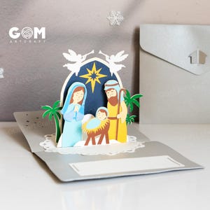 Pu&ograve; includere: Un biglietto di Natale pop-up con una scena della nativit&agrave; con Maria, Giuseppe e Ges&ugrave; bambino. Il biglietto &egrave; realizzato in carta azzurra, gialla e marrone, con una stella e angeli sullo sfondo. Il biglietto &egrave; aperto, rivelando la scena 3D.