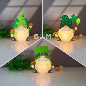 Puede incluir: Tres figuras luminosas de temática leprechaun verde con un diseño de trébol. Cada figura sostiene un pote de oro y lleva un sombrero verde con un trébol.