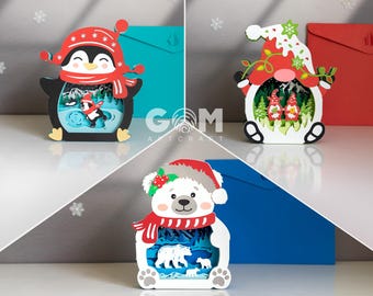 Puede incluir: Tres obras de arte navideñas en capas tridimensionales. Una presenta un pingüino con gorro y bufanda, otra un gnomo y la última un oso polar. Cada una tiene una escena invernal en su interior. El fondo incluye sobres azules y rojos.