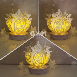 Könnte beinhalten: Drei weiße Papier-Schneeflockenlaternen mit filigranen Ausschnitten. Die Laternen werden von innen beleuchtet und erzeugen ein warmes Leuchten. Jede Laterne hat ein anderes Design: eine mit Kerzen, eine mit Glocken und eine mit einem Schneeflockenmuster.