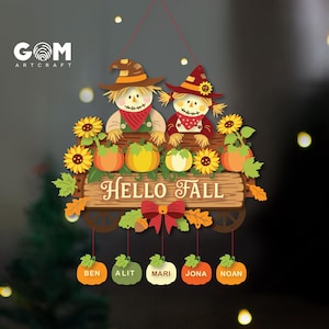 Può includere: Una targa decorativa da appendere con due spaventapasseri, girasoli e zucche su una tavola di legno con il testo HELLO FALL. Sotto, cinque piccole etichette a forma di zucca pendono da fili rossi, ciascuna con i nomi BEN, ALIT, MARI, JONA e NOAN.