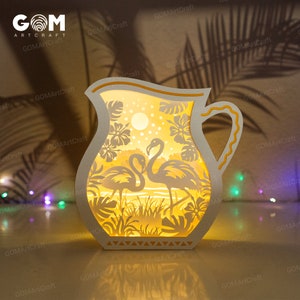 May include: A white wooden lamp shaped like a pitcher with a yellow handle. The lamp has a cut-out design of two flamingos and tropical plants. The lamp is lit and casting a warm glow.