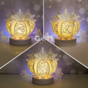 Könnte beinhalten: Weiße Papier-Schneeflockenlampe mit filigranen Ausschnitten, von innen beleuchtet, wodurch ein warmes Leuchten entsteht. Die Lampe steht auf einem weißen Sockel und ist von kleineren Schneeflocken umgeben.