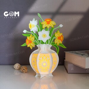 May include: A white paper vase with a floral design, lit from within, displaying paper daffodils in yellow, white, and orange. The vase is on a white surface. The text "GOM Art Craft" is visible.