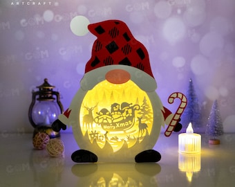Pode incluir: Um gnomo de Natal de madeira branco com um chapéu vermelho e preto xadrez segura um bastão de caramelo. A barriga do gnomo é uma janela iluminada com uma cena do Papai Noel em um trenó puxado por renas. O texto "Merry Xmas" é visível na janela.