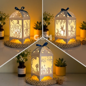 Puede incluir: Tres linternas de papel blancas con intrincados diseños recortados y luces LED. Las linternas presentan temas de Pascua, incluyendo conejos, huevos y las palabras "Feliz Pascua".