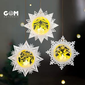 May include: Three white hanging star-shaped ornaments with intricate cut-out designs and glowing yellow centers. One features a nativity scene, one says Merry Christmas with an angel, and one shows two angels. The GOM ARTCRAFT logo is visible.