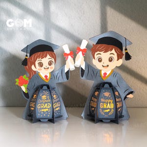 May include: Two graduation-themed paper cutouts. Each figure wears a graduation cap and gown, holding a diploma. The figures stand in front of a lantern with the words "Congrats Grad" and "Happy Grad". The figures are holding diplomas with red ribbons.