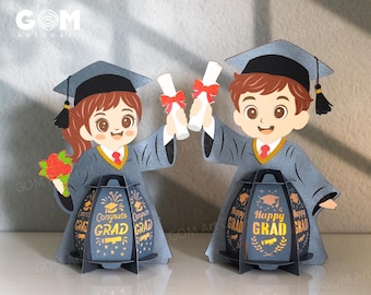 May include: Two graduation-themed paper cutouts. Each figure wears a graduation cap and gown, holding a diploma. The figures stand in front of a lantern with the words "Congrats Grad" and "Happy Grad". The figures are holding diplomas with red ribbons.