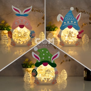 May include: Three Easter bunny night lights, each with a different colored hat. The bunny on the left is wearing a pink hat with white polka dots, the bunny in the middle is wearing a blue hat with white stars, and the bunny on the right is wearing a green hat with white stars. Each bunny is holding an Easter egg and has a small carrot in its hand.