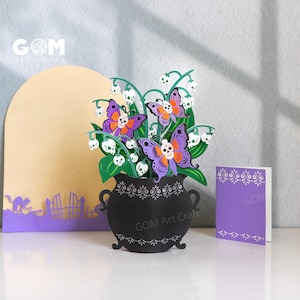Puede incluir: Una escena de manualidades de papel con un caldero negro con un arreglo floral de hojas verdes, flores blancas en forma de calavera y recortes de mariposas moradas y naranjas. Una tarjeta morada y un arco amarillo están en el fondo. El texto "GOM Art Craft" es visible.