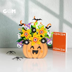 May include: A 3D paper craft centrepiece featuring a smiling orange pumpkin base filled with yellow star-shaped flowers, purple witch hats, white ghosts, and black bats. Two small black plastic spiders sit nearby, and a blank orange card stands beside it.
