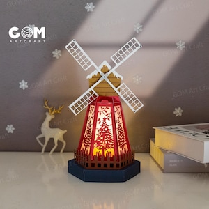 Puede incluir: Un molino de viento de papel rojo y blanco con una luz en el interior. El molino de viento está sobre una base de madera y tiene una valla alrededor de la parte inferior. La luz está encendida e ilumina el interior del molino de viento. El molino de viento está sobre una mesa con un libro al lado.