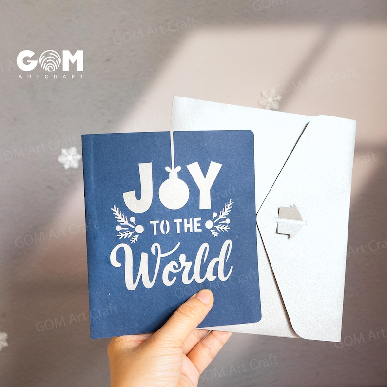 Pu&ograve; includere: Un biglietto di auguri blu navy con le parole "JOY TO THE WORLD" in scrittura bianca e una busta bianca. Il biglietto presenta un design festivo con un ornamento natalizio e ramoscelli.